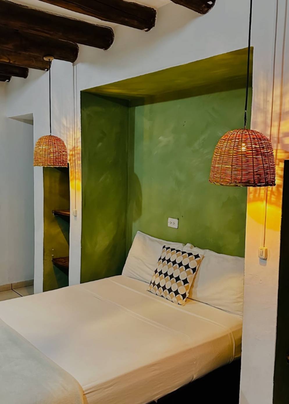 Habitación privada: cama blanca dentro de un nicho pintado de verde musgo, dos lámparas de mimbre encendidas y vigas de madera oscura.