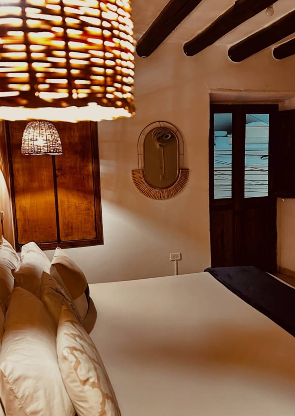 Habitación privada en luz cálida: lámpara de mimbre en primer plano, espejo de ratán en forma de sol, vigas oscuras y una ventana con postigos de madera.