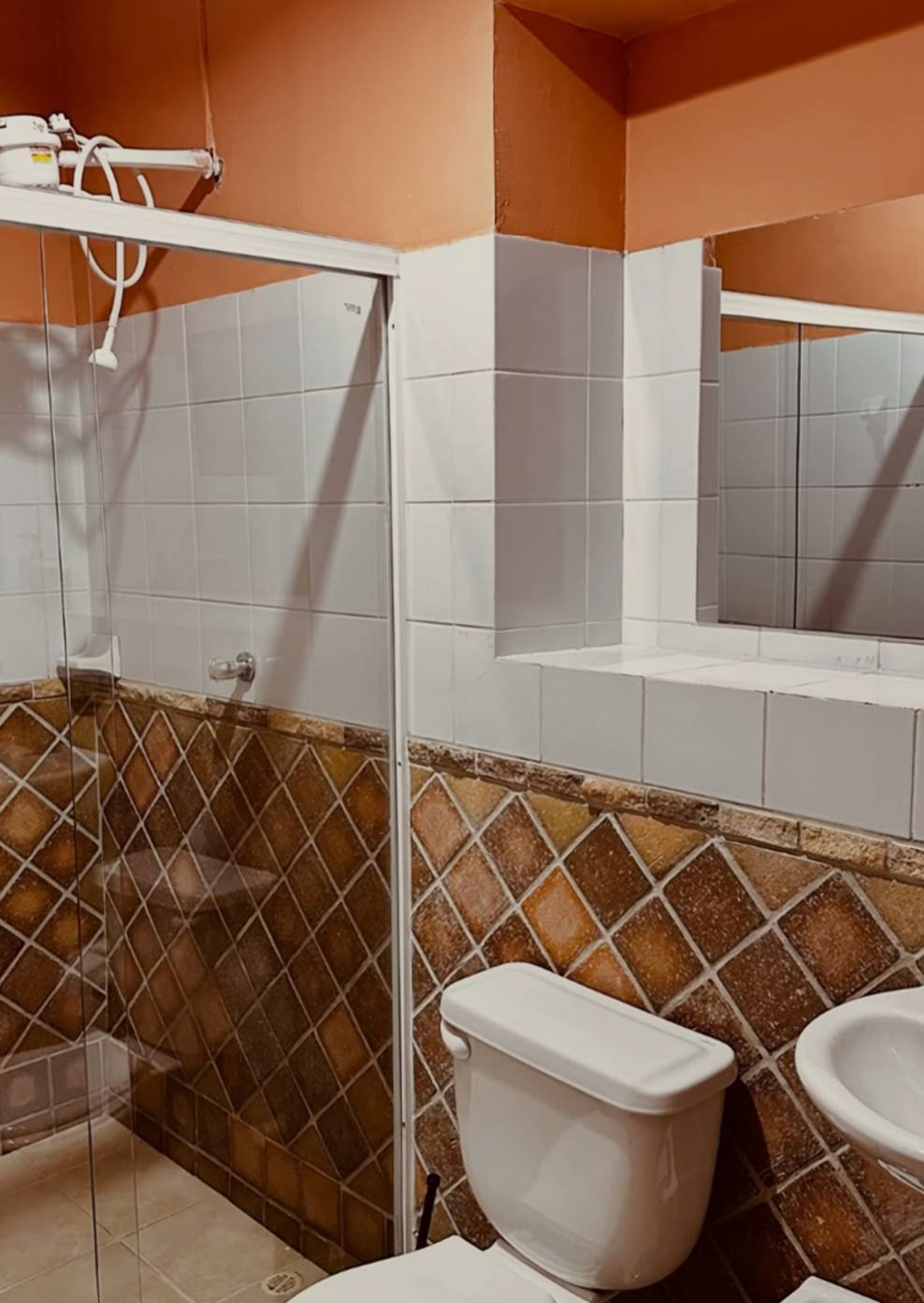 Baño privado con ducha de vidrio y calentador eléctrico, baldosas blancas y un zócalo de baldosa terracota en diagonal.