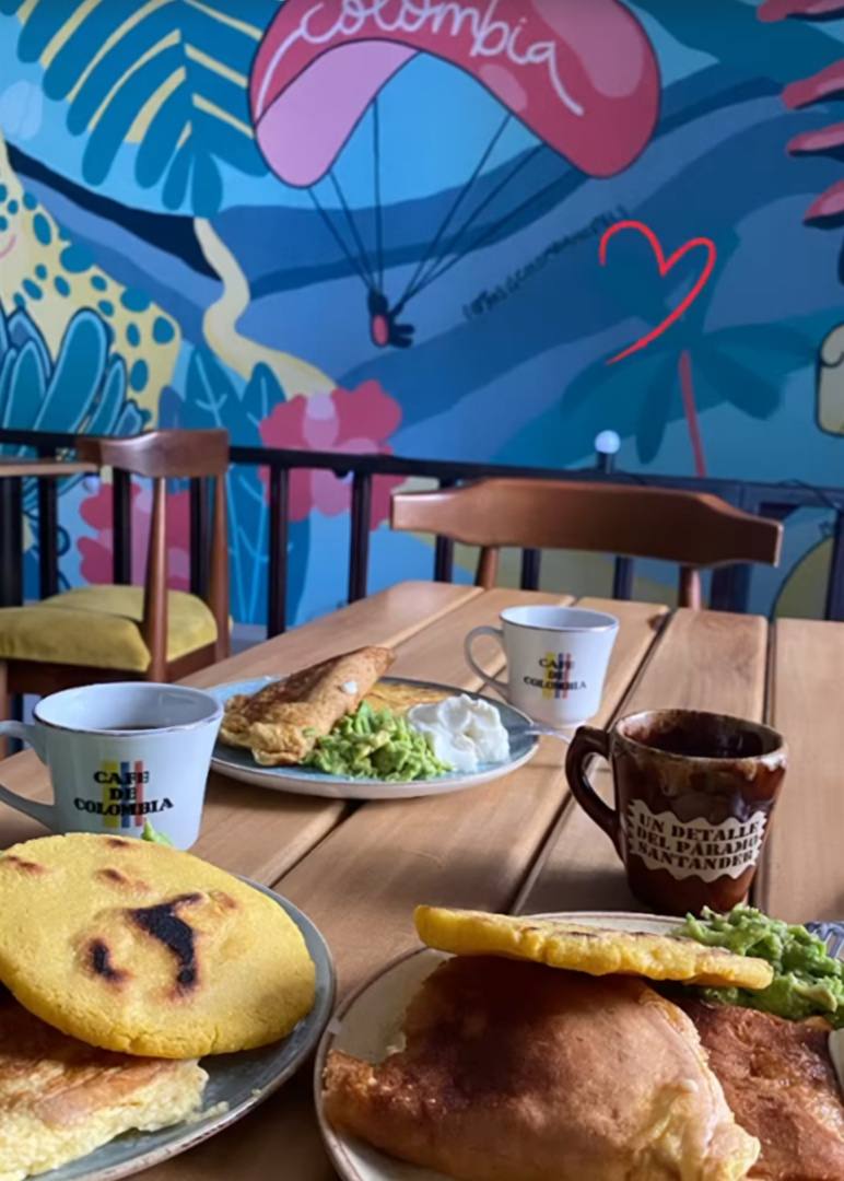 Mesa de desayuno con arepas, huevos, aguacate y tazas de café frente al mural de un parapente que dice «Colombia».